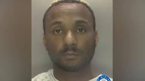 Walsh is perhaps earing a light-coloured medical gown in a police mugshot. He has short brown hair with a blonde tinge.