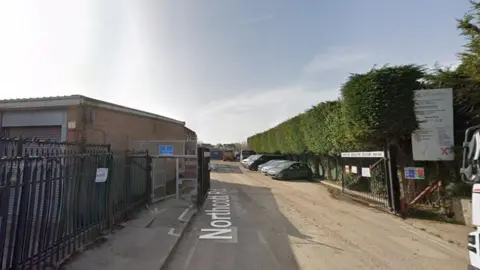 A road next to industrial buildings, with gates leading down the road. Hedges are on the right-hand side of the road and a row of cars are parked further down the road.