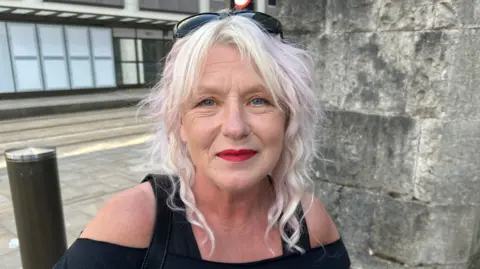 A woman with wavy white and pink hair wears a black top and sunglasses on her head