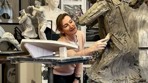 Amy Goodman is pictured in her workshop, using a tool on a statue in progress. Near her are figurines of two men on the prow of a boat, showing how part of the Coastal Forces memorial will look. Photographs of Royal Navy Wrens are positioned in front of her.