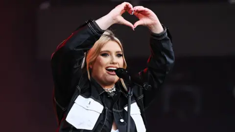Ella Henderson a woman with blonde hair who is performing at Radio 1's Big Weekend. She is wearing a denim jacket and is singing. 