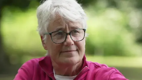 Ms Smith is wearing a pink top with glasses and has short grey hair. She is sitting outside.