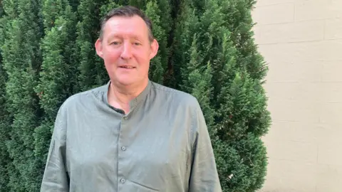 Dave Lee is standing in front of a green bush and a white wall. He is wearing a green shirt and has short hair. He is smiling at the camera.