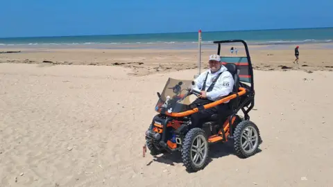 Submitted/George Pas George Pas wearing a grey hoodie and cap to commemorate the 80th anniversary of the D-Day landings. He sitting in his orange off-road wheelchair on Juno beach. It is a sunny day and the tide is out.