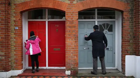 Getty Images An image of two unidentified political campaigners knocking at doors - there are two red doors to the left of frame where a woman in a pink coat and black hat is standing holding a clipboard, while on the left a man in a black coat and grey trousers is reaching up to the door knocker of a grey door