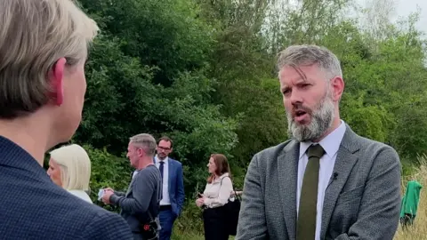 BBC Yorkshire's political editor James Vincent interviews Yvette Cooper MP at the site of the so-called Battle of Orgreave. He has a greying beard and is wearing a grey checked suit with a green tie. Ms Cooper has her back to the camera and is wearing a blue suit.