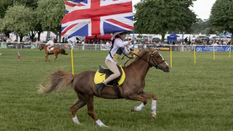A Gorman Photography A horse rider clutches a Union Jack flag as she rides along a course with people watching on in the distance at the Bath and West Show