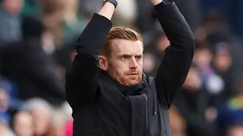Ed Still standing in the dugout with his arms above his head as he claps his hands