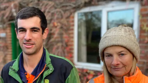 Luke Deal/BBC A man stands in the middle of two women. He is wearing a green farming overall and has dark short hair. The woman to his left is wearing a pink coat and wooly hat. The woman to his right is wearing an orange coat and cream wooly hat.