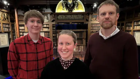 David, Lauren and Tony stand in an ornate old library surrounded by shelves of antique books. David is clean shaven with longish fair hair swept across his forehead. Lauren is smiling with fair hair tied back. Tony has a fair beard and short hair.