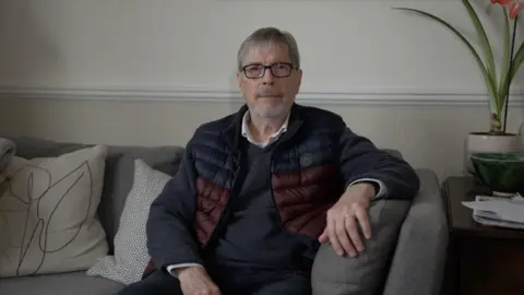 Nick Wells sits on a sofa. He is wearing a white shirt, blue sweater and a blue and maroon body warmer.