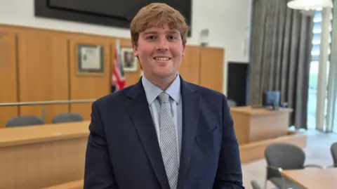Stuart Woodward/BBC Leader of Harlow Council, Dan Swords, stands in the council chamber of Harlow Council. He is wearing a navy blue suit, light blue shirt and patterned tie. He is smiling at the camera.