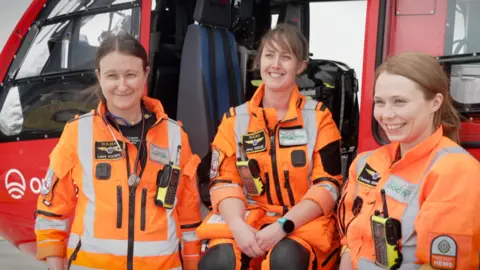 Three women side by side, in front of the red helicopter, wearing their orange uniforms, smiling at the person behind the camera.