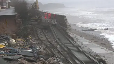 The picture shows a stretch of coastal railway that has been severely damaged by storm conditions, leaving the rails hanging and twisted. Chunks of ballast and earth have fallen away, exposing cables and debris. The sea is crashing in just a few metres away, and waves are rolling over the beach in the background. Three workers in high‑visibility clothing are standing on what remains of the wall, next to a digger that appears to be part of the emergency repair effort.