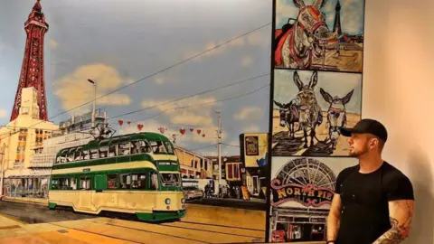 Adam Salisbury Artist Adam Salisbury wearing a black baseball cap and black t-shirt standing by his huge painting of a green and white tram on Blackpool Promenade with Blackpool on a wall at Blackpool Victoria Hospital. He is looking at the artwork. 