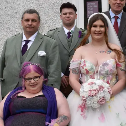 Alysha McLean Alysha McLean, wearing a wedding dress, standing next to her mother, who has dyed pink hair and is wearing dark clothing.