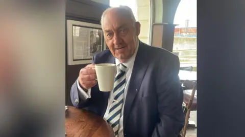 A man with grey hair wearing a blazer, tie and white shirt sitting at a table while holding up a coffee mug.