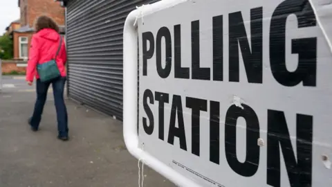A woman walking in front of a shuttered shop, with a polling station sign in the foreground 