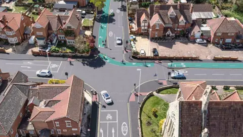 Gloucestershire County Council A computer-generated image of cars using a junction surrounded by Edwardian or Victorian houses which has a green cycle path going through it. 