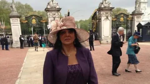 Savitri Cameron A woman in a purple dress and purple blazer looks at the camera in front of the gates of Buckingham Palace. She wears her black hair down, with silver dangly earrings, a silver necklace, and blush pink-brimmed hat with floral detail