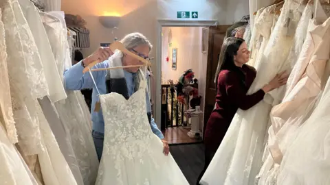 A woman with long brown hair sorts through hanging wedding dresses while a woman with a blue jumper and scarf holds up a floral white dress