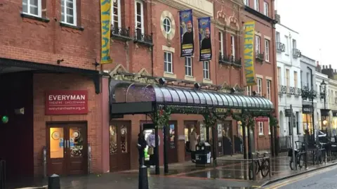 The exterior of Cheltenham's Everyman Theatre on a rainy day. It is a brick building with a glass canopy out the front on a Regency style town centre street. There are people sheltering from the rain beneath the canopies.