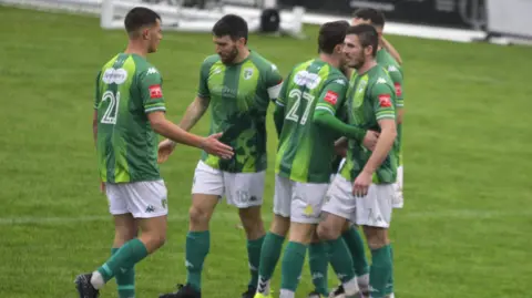 Fran Torode Six Guernsey FC players celebrate with each other after scoring a goal. All are wearing the football club's green home kit with white shorts and green socks.