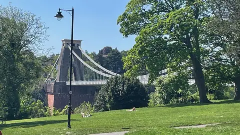 Two people are seen in the distance sunbathing by the Clifton Suspension Bridge on a sunny day