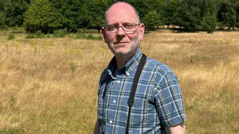 BBC / Gavin Kermack A man, blad on top but with grey hair on the sides of his head, standing in a grassland field with trees in the background. It is clearly a summer's day. He is wearing thin-rimmed glasses and a short-sleeved check shirt and has something out of frame hanging around his neck.