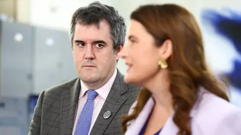 Getty Images A man with grey hair and a purple tie looks over as a woman in a purple suit speaks