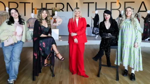 Four women are in a row. Left is Hope Macauley who has long dark hair and is wearing jeans and a chunky, light coloured cardigan. She is standing. Next is Sara O'Neill who is sitting on a high stool, wearing a long dark dress and long jacket and dark sandals. She has long fair hair and is smiling. In the middle is Judith Owens of Titanic Belfast who is wearing a red trouser suit, and standing. She has long blonde hair. Beside her, is Gráinne Maher who is sitting on a stool, wearing a dark top and trousers and a dark beret. She has long dark hair and is wearing bright red lipstick. Finally, to the right of the picture is Amy Anderson who is wearing a long pale green and purple striped dress and dark flat shoes with white socks or rights. She has blonde hair. The women are in front of a row of of manikins.