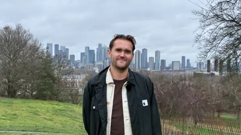 A young white man with brown hair, dressed in brown tshirt, white shirt and black jacket smiles at the camera with a backdrop of trees and skyscrapers