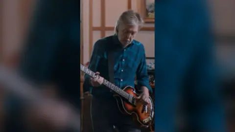 Paul McCartney wearing a blue shirt and playing a brown bass.