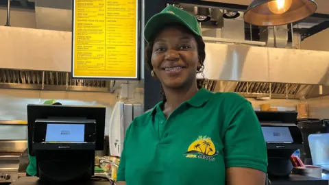 Woman wearing cap and polo shirt smiling with kitchen and electronic menu behind her.