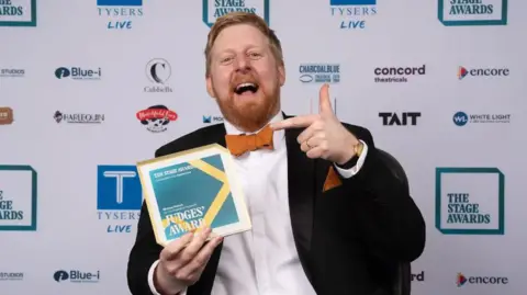 Michael Campbell is wearing a black suit, white shirt and orange bow tie. 
He is seen holding the Judges' Award from the Stage Theatre Awards.