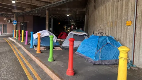 Four tents are visible by the side of a road leading into an indoor car park.