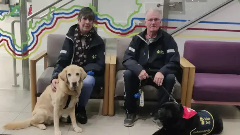 Stuart Geddes A wide shot of Marylou and Peter sitting on a sofa. On the floor in front are the two dogs.
