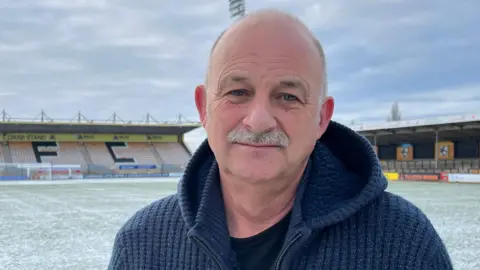 Ian Darler is in the middle. He has a grey moustache and is wearing a blue jumper and a black T-shirt. He is stood on a football pitch which has a light layer of snow covering it.
