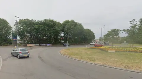 The roundabout in Burnham-on-Sea at the junction between the B3140 and Love Lane. There are four exits and a green island in the middle, with trees and shrubs planted on it.