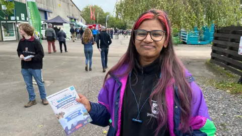 Azra Anzar stands with a programme for the Dairy Show, which she is handing out. Behind her people stream into the show.