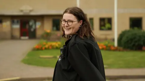 Ministry of Justice Hollie Peabody pictured outside the prison where they work in their black prison officer uniform. They look over their left shoulder to smile at the camera, surrounded by a neat lawn and a flower bed planted with orange and yellow flowers. 