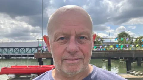 BBC / Lara King Malcolm Pearce wearing a blue top smiling at the camera. He is standing in front of a red boat and water with some bunting in the background