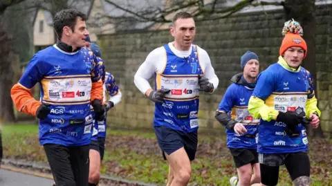 Danny Lawson/PA Wire Kevin Sinfield, triathlete Jonny Brownlee and boxer Josh Warrington are wearing blue race jerseys with printed sponsor logos jog along a wet roadside during an organized event