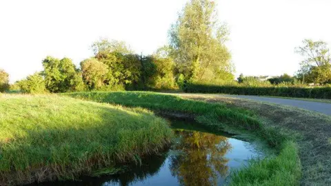 Ruth Sharville / Geograph.org.uk A field of grass and road separated by a body of water.