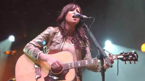 KT Tunstall sings while playing guitar. She has long brown hair and is wearing a brown leather jacket.