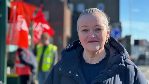 Debbie Hallsall, who has blonde hair that has been brushed back and is wearing a blue padded winter coat. Bus drivers in hi-vis holding red unite flags are standing at a green bus stop behind her.