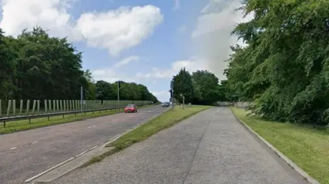 A grey road with a red car on it. Green trees and bushes line the road.