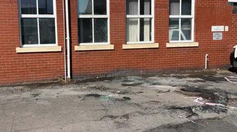 The photo shows the side of a red brick building which has four windows. The tarmac beneath it has holes in and appears to have been scorched. In some places it has turned black. 
