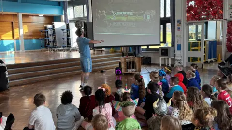 Ash Randall balances a ball on his head in front of a school assembly. The children are seated on the floor watching him.