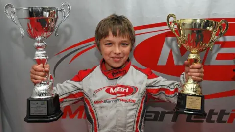Mick Barrett Racing Childhood photo of Lando Norris. He has brown short hair and is wearing a racing suit. He is holding two trophies - one silver, and one gold